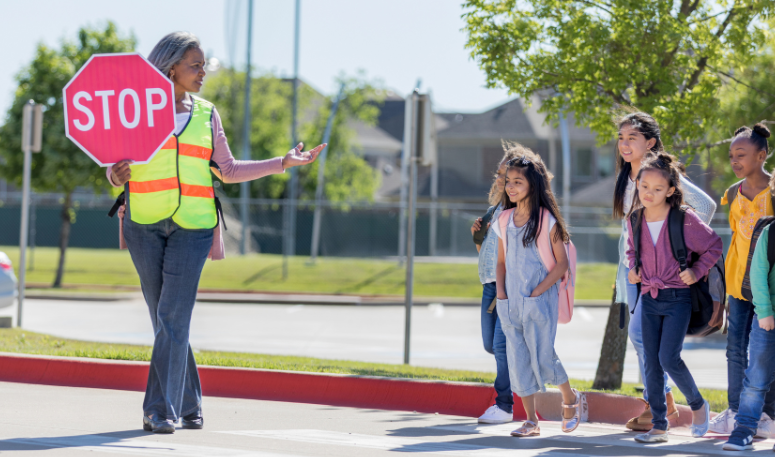 Children wait at a crosswalk as a crossing guard signals for them to cross the street.