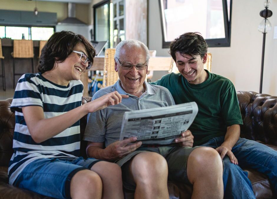An older man sitting on a couch reading a newspaper while two younger people smile and point at the page.