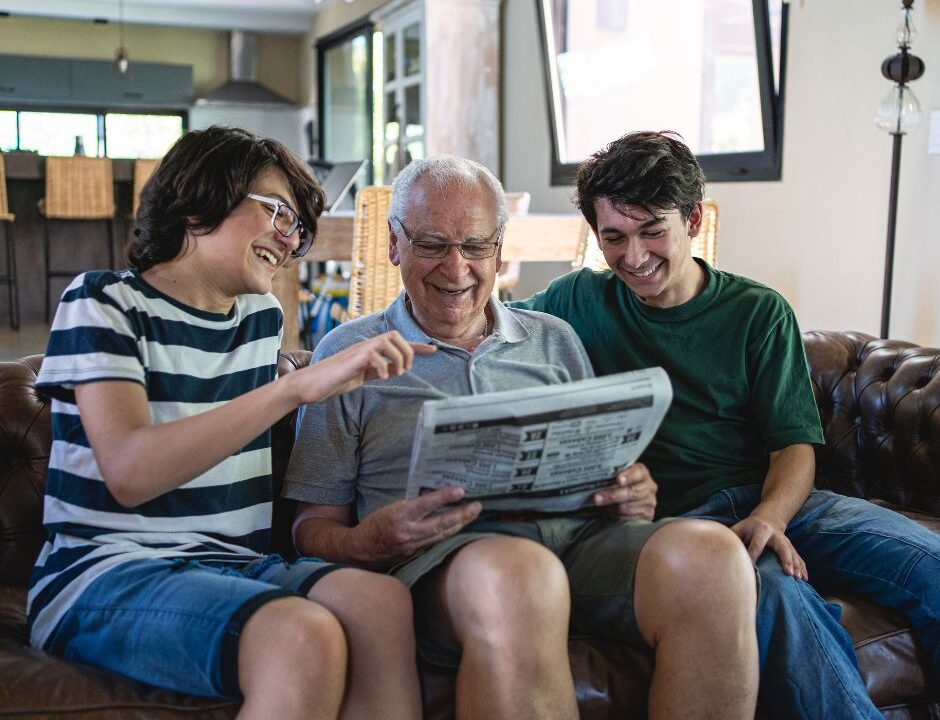 An older man sitting on a couch reading a newspaper while two younger people smile and point at the page.